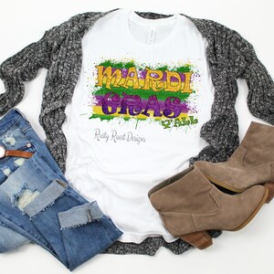 May include: White t-shirt with a colorful Mardi Gras graphic design that says "Mardi Gras Y'all" with green, purple, and gold paint splatters. The shirt is laying on a gray cardigan, blue jeans, and brown boots.
