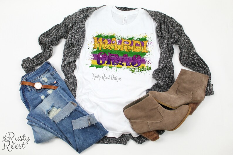 May include: White t-shirt with a colorful Mardi Gras graphic design that says "Mardi Gras Y'all" with green, purple, and gold paint splatters. The shirt is laying on a gray cardigan, blue jeans, and brown boots.