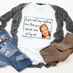 May include: White t-shirt with a vintage-style graphic featuring a woman and the text "If you can't say anything nice, then you should come sit by me!"