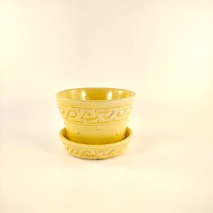 May include: A pale yellow ceramic planter with a matching saucer. The planter has a cylindrical shape with a decorative Greek key pattern around the top and a row of small dots. The saucer also features the Greek key design. The set is suitable for small plants.
