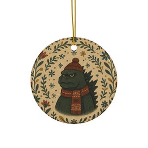 Può includere: Ornamento natalizio rotondo in ceramica. Presenta un personaggio di Godzilla verde con cappello e sciarpa rossi. L'ornamento ha un bordo floreale con fiocchi di neve su sfondo beige e un cordino dorato per appenderlo.