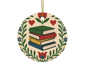 May include: Round ceramic ornament with a stack of books in green, red, yellow, and blue. The ornament has a cream background with a floral and heart design. A gold string is attached for hanging.