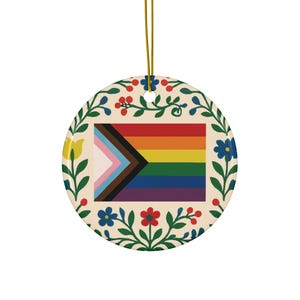 May include: Circular ceramic ornament displaying the Progress Pride flag, incorporating a rainbow design and a chevron in black, brown, light blue, pink, and white. The ornament is framed by a floral border with red, blue, and yellow flowers and green leaves. A gold hanging string is attached.