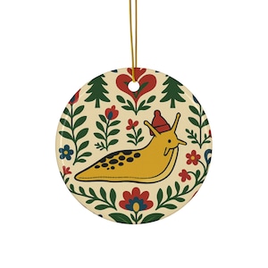 Puede incluir: Adorno redondo de cerámica con un caracol amarillo con un gorro rojo, rodeado de diseños florales y de árboles. El adorno tiene una cuerda dorada para colgar. El fondo es crema.