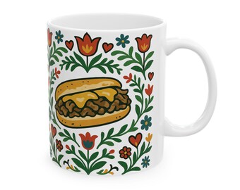 May include: White ceramic mug featuring a Philly cheesesteak illustration surrounded by a floral design with red, orange, and blue flowers. The mug has a curved handle and is suitable for hot or cold beverages.
