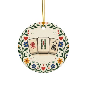 May include: A circular ceramic ornament displaying three Mahjong tiles with Chinese characters. The tiles are set against a floral design with red hearts, blue and yellow flowers, and green leaves. A gold string is attached for hanging.