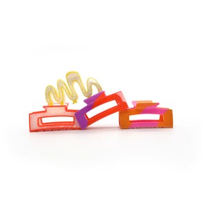 Puede incluir: Dos pinzas para el pelo de plástico de colores. Una pinza es roja, blanca y morada con un diseño de espiral amarillo. La otra pinza es rosa, roja y naranja.