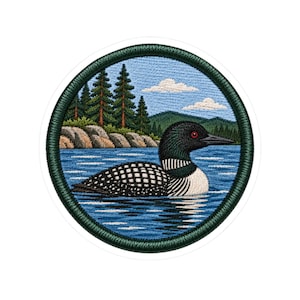 May include: A circular embroidered patch featuring a detailed scene of a black and white loon swimming on blue water. The background displays a rocky shoreline, a dense forest of green pine trees, distant mountains, and a light blue sky with clouds.