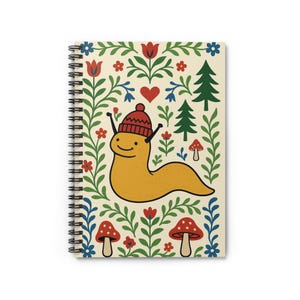 Könnte beinhalten: Spiralgebundenes Notizbuch mit einer skurrilen Illustration einer lächelnden gelben Schnecke mit roter Strickmütze. Das Design umfasst einen floralen Rand mit roten Pilzen, Blumen und grünen Blättern. Der Hintergrund ist cremefarben.