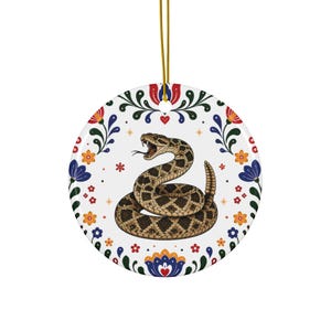 Könnte beinhalten: Runder Keramikschmuck mit einer aufgerollten Klapperschlange mit offenem Maul, die braune und beige Rautenmuster aufweist. Der Schmuck ist von einem floralen Rand mit roten, orangefarbenen, blauen und grünen Akzenten und einer goldenen Aufhängeschnur umgeben.