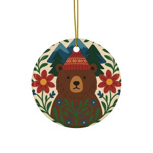 May include: Round ceramic ornament featuring a brown bear wearing a red knit hat. The bear is surrounded by red flowers, green leaves, and blue flowers. The background includes mountains and trees. A gold string is attached for hanging.
