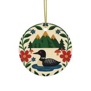 May include: Round ceramic ornament featuring a stylized illustration of a loon swimming in a lake, with mountains and trees in the background. The ornament is decorated with red flowers, blue flowers, and green foliage. A gold string is attached for hanging.