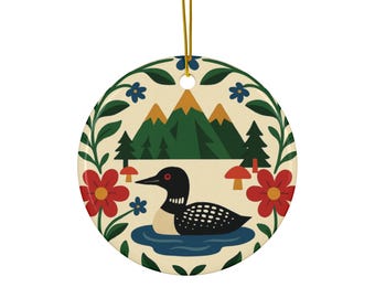 Könnte beinhalten: Runder Keramikschmuck mit einer stilisierten Illustration eines Eistauchers, der in einem See schwimmt, mit Bergen und Bäumen im Hintergrund. Der Schmuck ist mit roten Blumen, blauen Blumen und grünem Laub verziert. Ein goldener Faden ist zum Aufhängen befestigt.