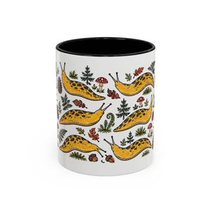 May include: A white ceramic mug with a black interior and rim, featuring a repeating pattern of yellow slugs, small red and white mushrooms, green ferns, pine trees, brown acorns, and various autumn-colored leaves scattered across the surface.