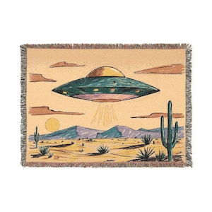 May include: A woven tapestry depicting a cartoon drawing of a flying saucer hovering over a desert landscape with cacti. The saucer is green and purple, and the desert is beige and brown.