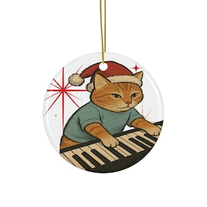 May include: A round Christmas ornament featuring a cartoon orange tabby cat wearing a Santa hat and a teal shirt, playing a piano. The ornament has a gold string for hanging and a red starburst design on a white background.
