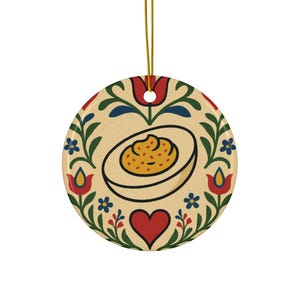 May include: A round ceramic ornament featuring a deviled egg design. The egg is surrounded by a floral pattern with red tulips, blue flowers, and green leaves. A gold string is attached for hanging.