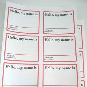 以下が含まれることがあります： 赤い枠で囲まれた6つのボックスがある印刷可能なワークシート。各ボックスには「こんにちは、私の名前は」というテキストと、名前を書くためのスペースがあります。各ボックスの下には「私の性別の代名詞は」というテキストと、代名詞を書くためのスペースがあります。
