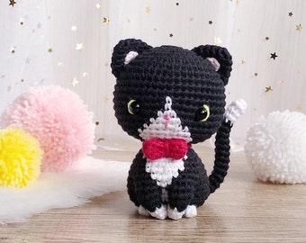 May include: A handmade black and white crochet cat with a pink bow tie and yellow eyes. The cat has a white bib and white accents on its ears and tail. The background includes pink and white pom-poms and a white surface.