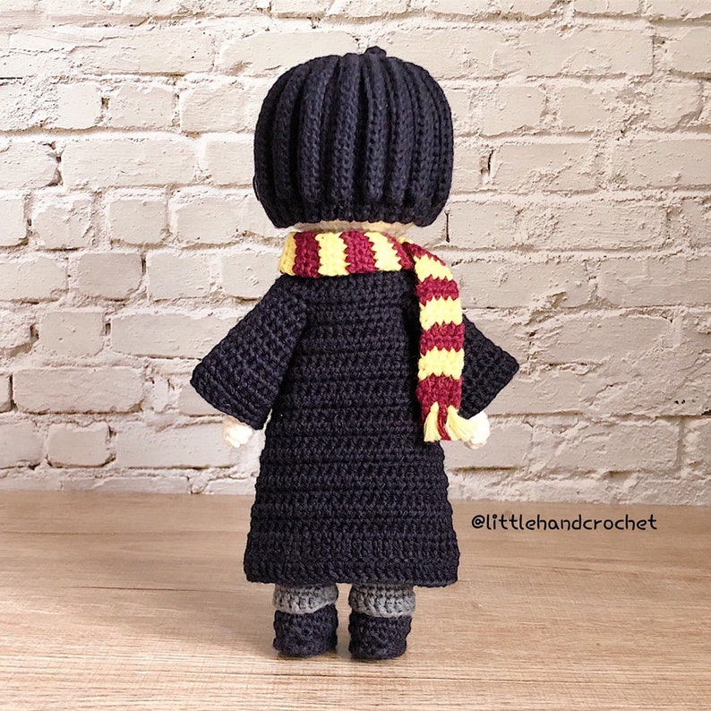 May include: A crocheted doll wearing a black robe, a black hat, and a red and yellow scarf. The doll is standing on a wooden surface.