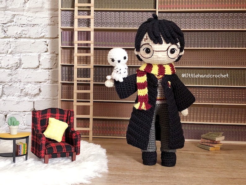 May include: Crocheted amigurumi doll of Harry Potter wearing a black robe, a red and gold scarf, and round glasses. The doll is holding a small white crocheted owl. The doll is standing in front of a bookshelf.