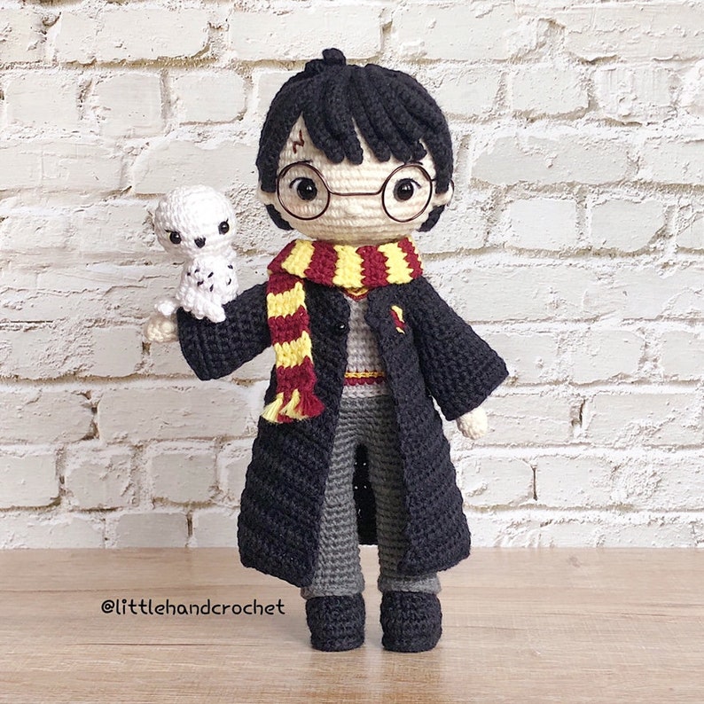 May include: Crocheted Harry Potter doll with a black robe, a yellow and red scarf, and round glasses. The doll is holding a white crocheted owl.
