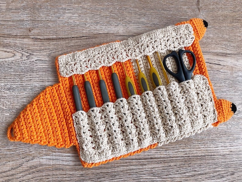 May include: A crocheted fox-shaped case with a beige interior and orange exterior. The case has eight slots for holding knitting needles and a separate slot for scissors. The case is open and the needles and scissors are visible.