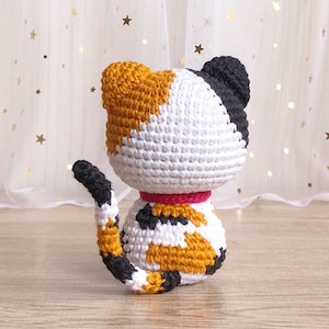 May include: A crocheted calico cat toy with black, white, and orange yarn. The cat is sitting with its tail curled around its body. It is wearing a pink collar.