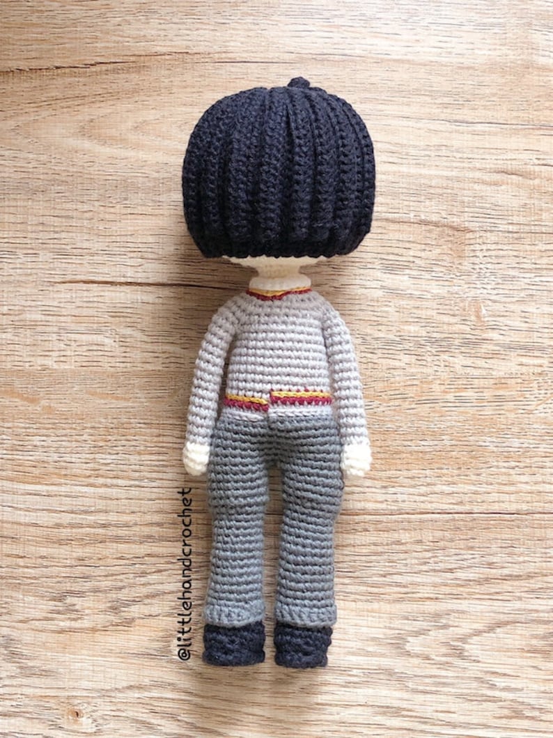 May include: A crocheted doll with black hair, a gray sweater, a red and gold belt, gray pants, and black shoes.