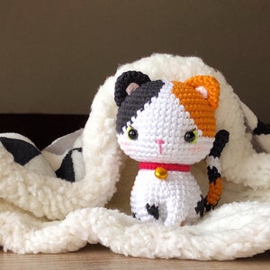 May include: A crocheted calico cat toy with black, white, and orange fur. The cat has a red collar with a gold bell. The cat is sitting on a white fluffy blanket.