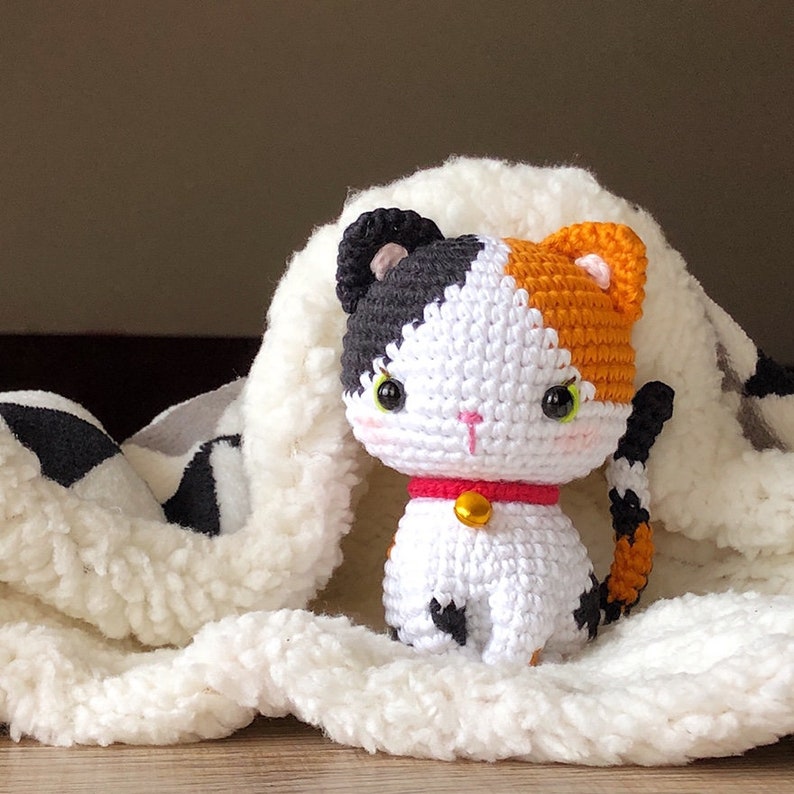 May include: A crocheted calico cat toy with black, white, and orange fur. The cat has a red collar with a gold bell. The cat is sitting on a white fluffy blanket.