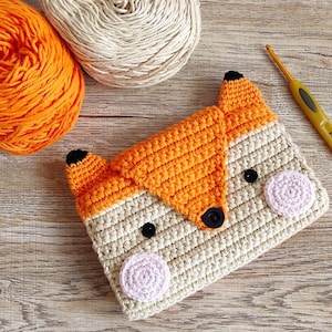 May include: A crocheted fox-shaped pouch with a beige body and orange fox face. The pouch has black eyes, a black button nose, and pink felt cheeks.