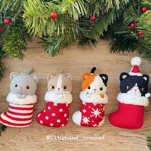 Könnte beinhalten: Vier gehäkelte Katzenornamente in Weihnachtsstrümpfen. Die Katzen sind grau, hellbraun, orange und schwarz, jede mit einem einzigartigen Design. Die Strümpfe sind rot und weiß, mit verschiedenen Mustern. Das Bild wird von Tannenzweigen und roten Beeren umrahmt.