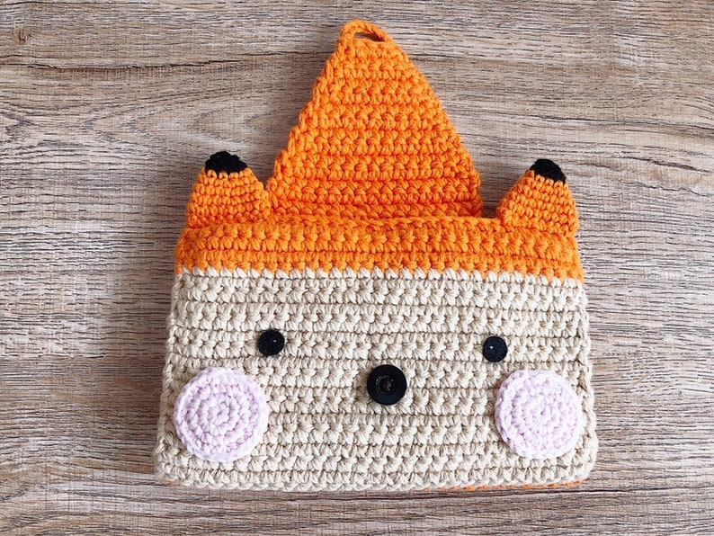 May include: A crocheted fox-shaped pouch with orange ears and a beige body. The pouch has black eyes and a pink nose.