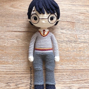 May include: A crocheted Harry Potter doll wearing a grey sweater, black pants, and round glasses. The doll has black hair and a lightning bolt scar on his forehead.