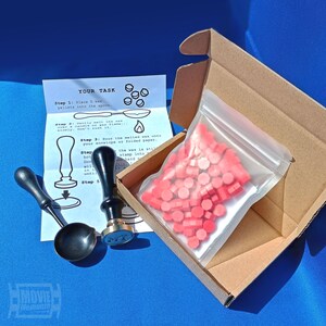 May include: A wax seal kit with a black spoon, stamp, and pink wax pellets in a clear bag inside a cardboard box. Instructions are printed on a white paper. The text on the paper reads "YOUR TASK".