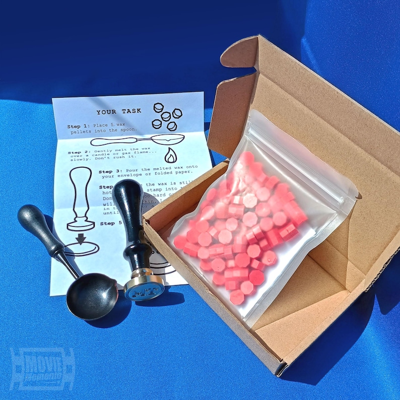 May include: A wax seal kit with a black spoon, stamp, and pink wax pellets in a clear bag inside a cardboard box. Instructions are printed on a white paper. The text on the paper reads "YOUR TASK".