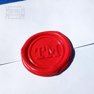 May include: A bright red wax seal with the letters "TM" embossed in the center. The seal is circular and sits on a white surface, likely an envelope. The image is well-lit, highlighting the texture and color of the wax.
