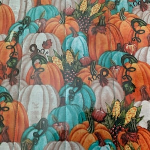 May include: A quilted fabric featuring a repeating pattern of pumpkins in shades of orange, teal, and white. The pumpkins have green vines and are interspersed with corn and apples, creating a fall harvest theme.