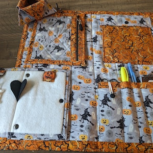 May include: A Halloween-themed organizer with multiple pockets and compartments. The fabric features a gray background with pumpkins, witches, and ghosts. Orange and black accents frame the organizer, and a small pumpkin patch is visible.
