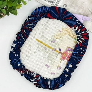 May include: A cross-stitch project in a navy blue fabric frame with a red and white starburst pattern. The cross-stitch depicts a figure in a red and purple outfit holding a yellow staff.