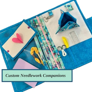 May include: A blue needlework organizer with multiple pockets and compartments. It features a clear vinyl pocket with a pink heart, a small pouch, and a ruler. The text "Custom Needlework Companions" is displayed.