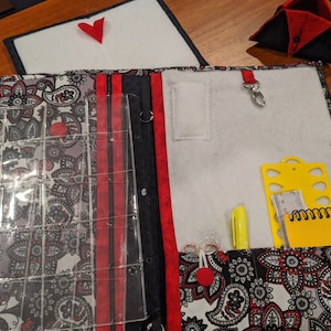 May include: A black and white paisley fabric organizer with red accents. The organizer has a clear plastic pocket with multiple compartments, a zippered pocket, and a small pocket with a key ring. The organizer is open and shows a yellow ruler, a yellow pencil, and a yellow notepad.