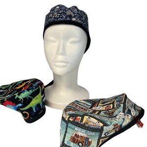 Puede incluir: Tres gorros quirúrgicos con diferentes patrones. Un gorro tiene un fondo negro con dinosaurios coloridos, otro tiene un fondo azul con un tema de viaje vintage y el tercero tiene un fondo negro con un diseño de calavera y huesos cruzados.