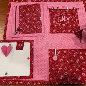 May include: A pink and red fabric organizer with multiple pockets and compartments. The organizer has a white felt insert with a heart and a key chain. The organizer also has a clear plastic pocket and a pocket with the initials "JMW" embroidered on it.