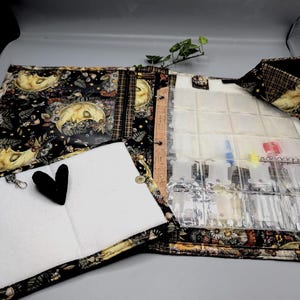 May include: Open, fabric organizer with a black and gold celestial print featuring moons and stars. The organizer has clear pockets for vials, a white padded section, and a black heart charm. It is designed to hold essential oils.