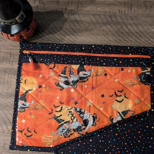 May include: A quilted zippered pouch with a black background and colorful polka dots. The front features a whimsical Halloween design with gnomes riding broomsticks against a backdrop of a full moon, stars, and bats.
