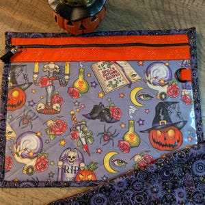 May include: A purple quilted zippered pouch with a clear vinyl front. The pouch features a Halloween-themed print with images of pumpkins, skulls, potion bottles, stars, and a book that says "Spells, Potions & Recipes".