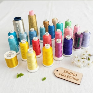 Puede incluir: Una colección de coloridas bobinas de hilo en varios tonos de azul, rosa, amarillo, rojo y morado. También hay una etiqueta de madera con el texto "SPRING COLLECTION". Los hilos están dispuestos sobre una superficie blanca.