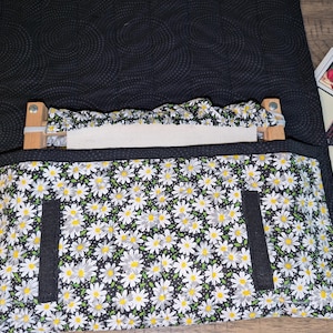 Puede incluir: Una bolsa de proyecto de tela negra con estampado de margaritas blancas. La bolsa tiene una parte superior con cremallera y dos bolsillos en la parte delantera. La bolsa está abierta y hay un aro de bordado de madera en el interior.